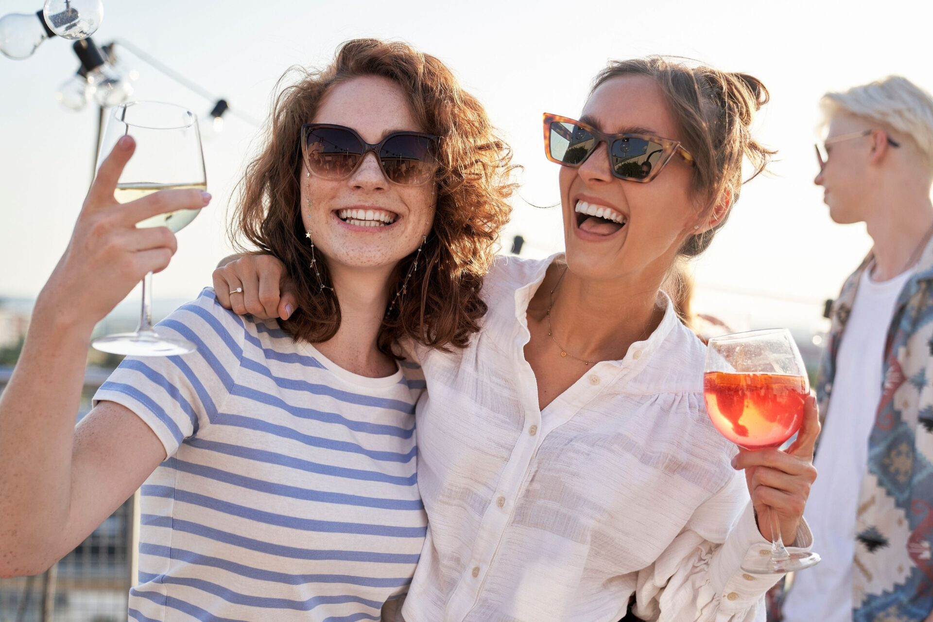 Zwei lachende Frauen mit Sonnenbrille, Wein und einem orangen Drink in der Hand, im Hintergrund ein junger Mann