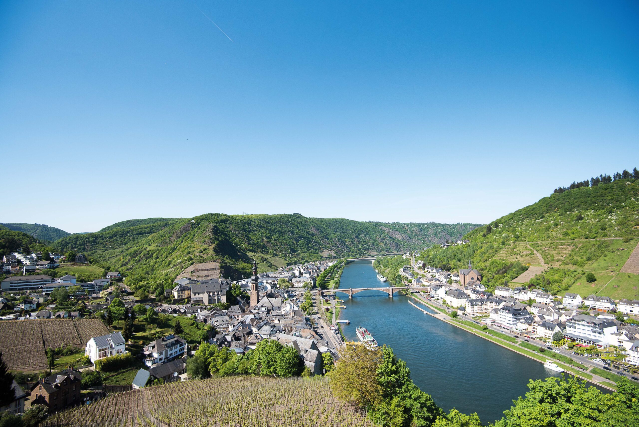 Ein weiter Blick über die Mosellandschaft bei Cochem. Im oberen Teil des Bildes sieht man einen blauen Himmel, im unteren Teil sieht man die Mosel, die sich durch die Stadt Cochem erstreckt.