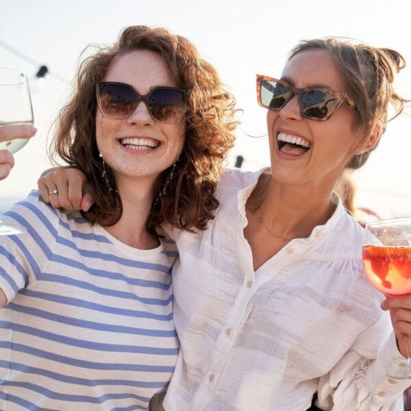 Two smiling women with sunglasses, wine and an orange drink in their hands, a young man in the background