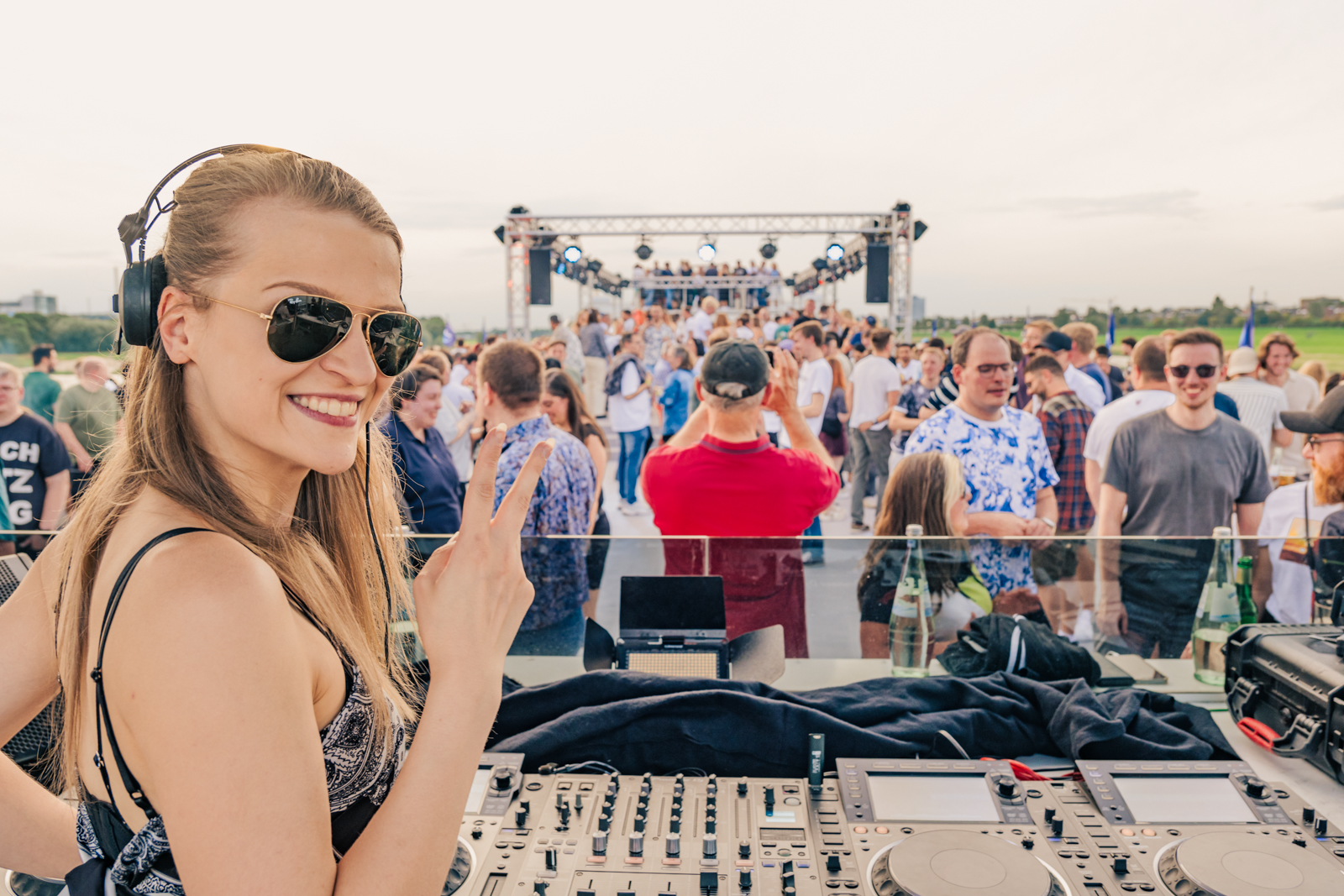 DJane Sintica mit Sonnenbrille un Kopfhörern vor ihrem DJ-Pult zeigt das Peace Zeichen in die Kamera. Im Hintergrund feiernde Leute auf dem Freideck.