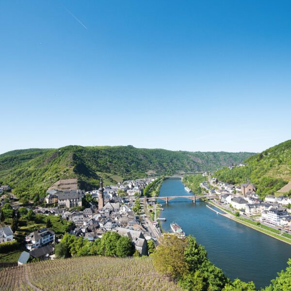 Ein weiter Blick über die Mosellandschaft bei Cochem. Im oberen Teil des Bildes sieht man einen blauen Himmel, im unteren Teil sieht man die Mosel, die sich durch die Stadt Cochem erstreckt.